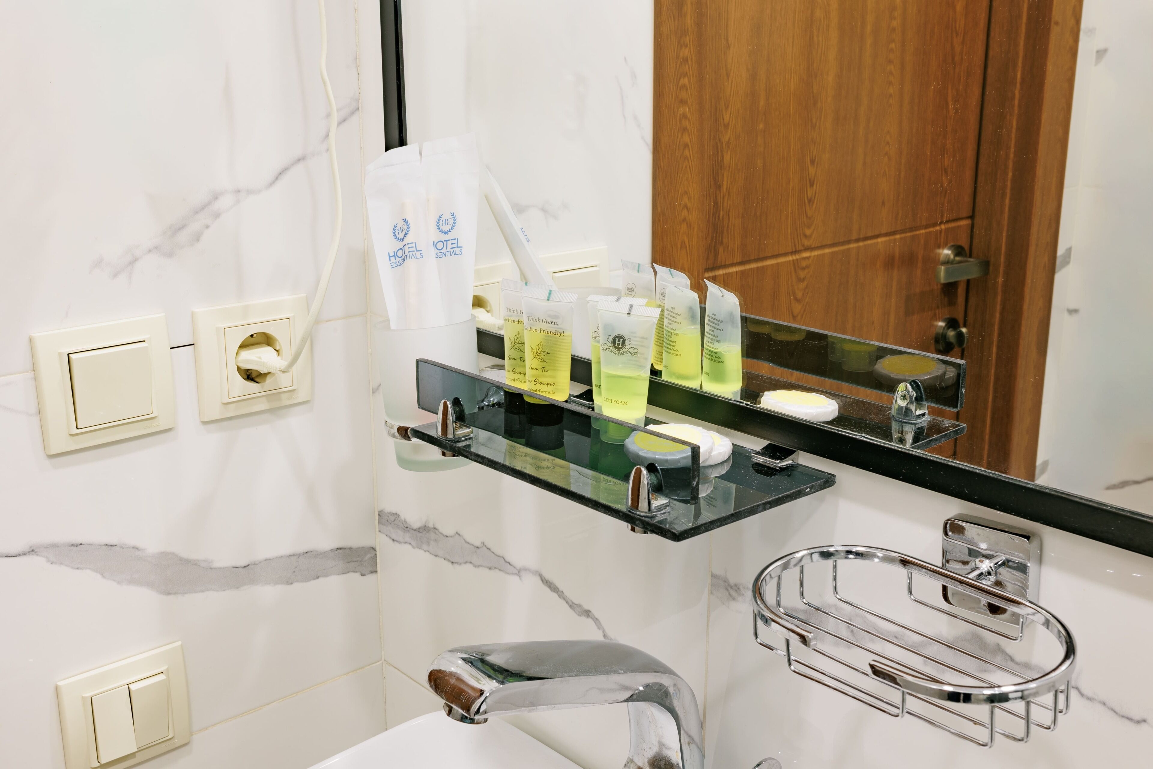 A bathroom with a marble wall, a glass shelf with toiletries, a wooden door, and a chrome sink.