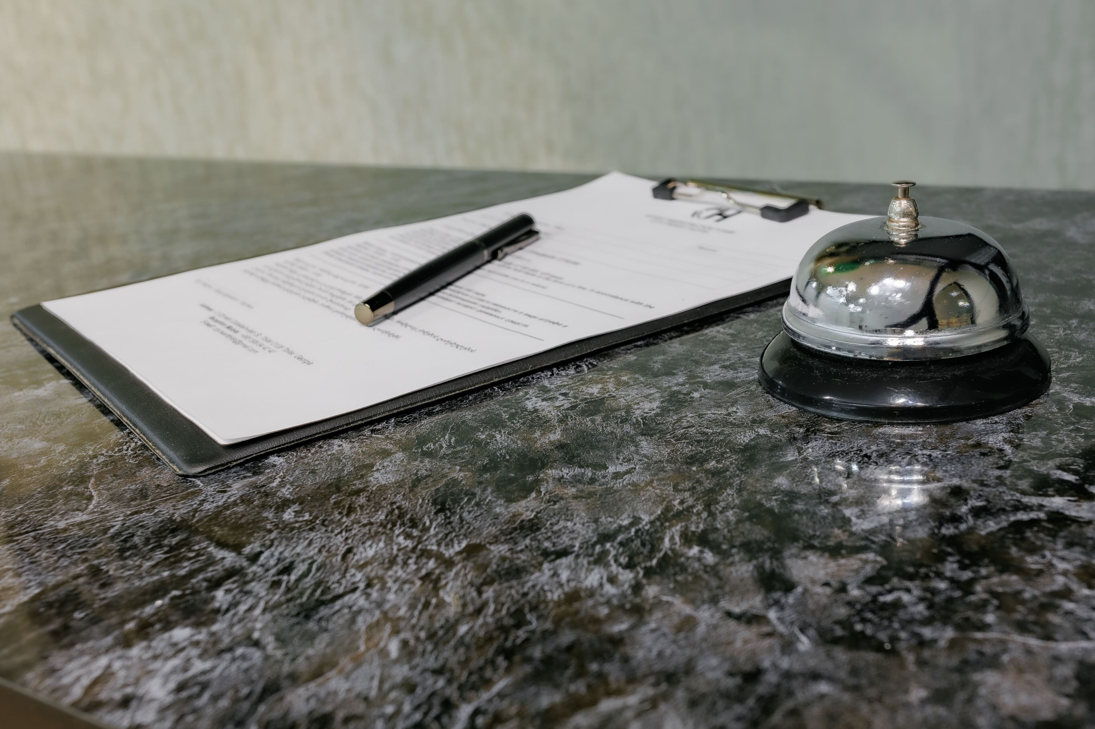 A clipboard with a pen on a marble surface, a small bell, and a document.