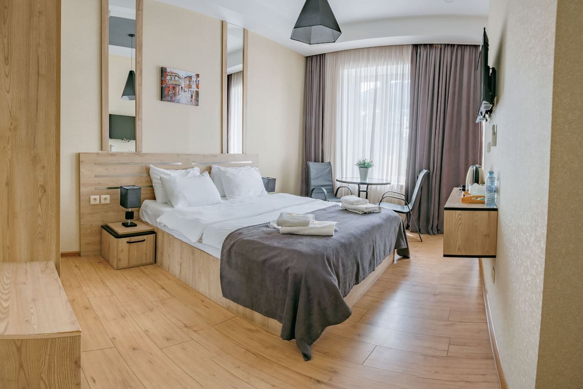 A hotel room with a wooden floor, a bed with white linens, a bedside table with a lamp, a chair, a desk with a plant, and a TV mounted on the wall.