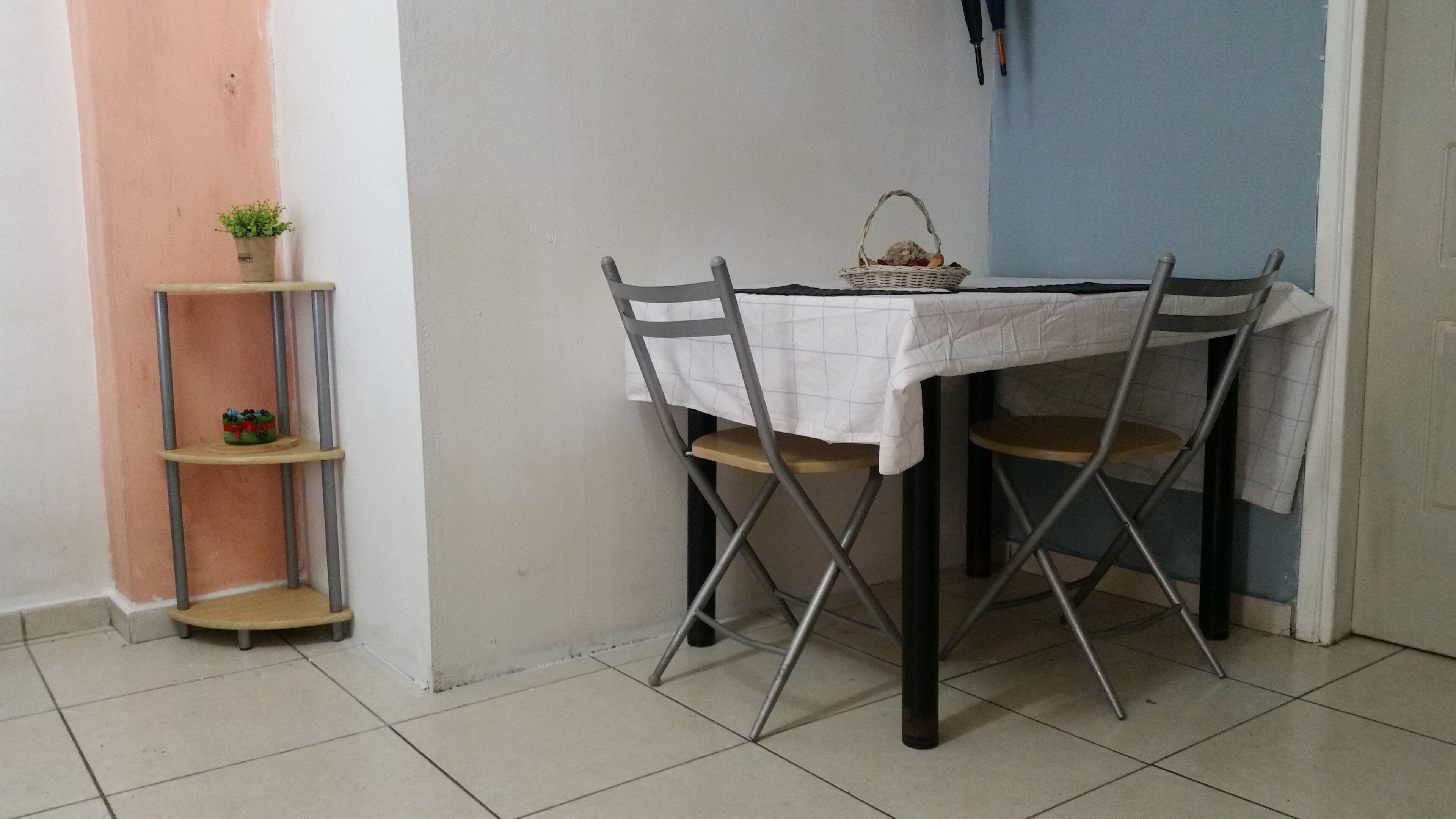 Un comedor con una mesa dispuesta para dos, una silla de metal, un estante con planta en maceta y una cesta sobre la mesa.