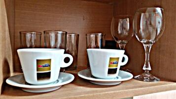 Dos tazas blancas con un logotipo sobre un estante de madera, acompañadas por vasos transparentes y una copa de vino.
