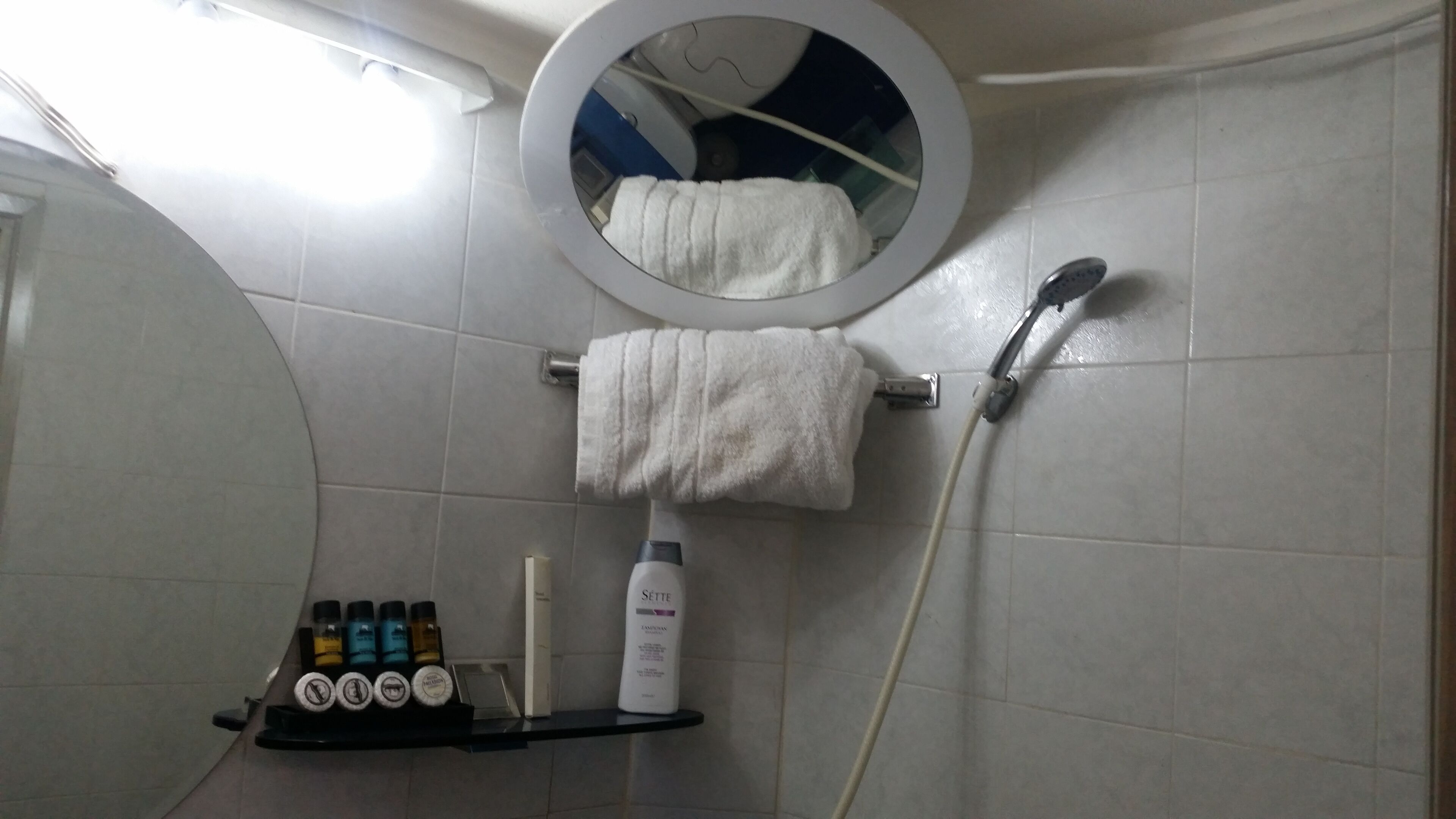 Un baño con un espejo redondo, un perchero con una toalla blanca, un cabezal de ducha y un estante con artículos de aseo.