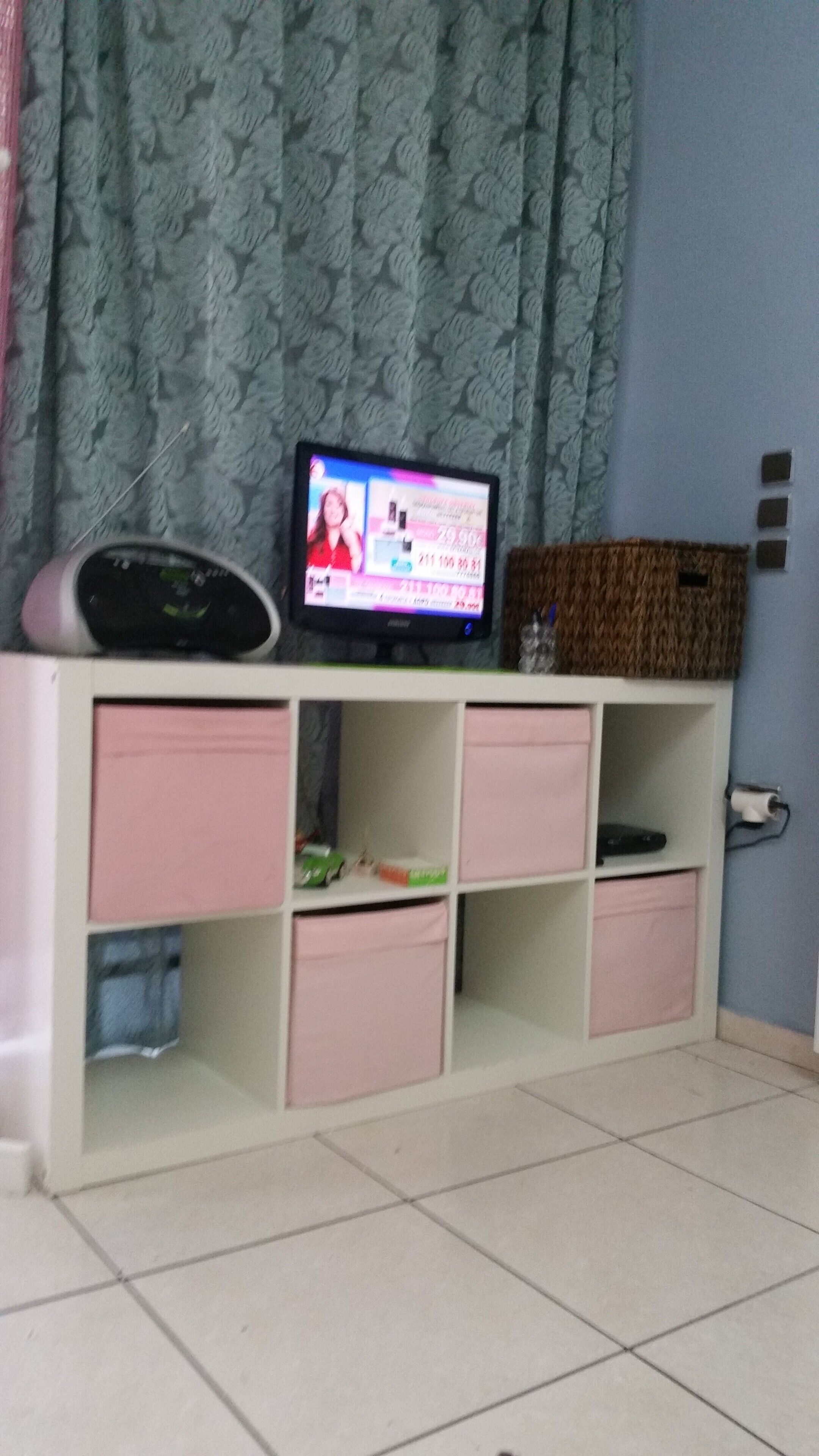 Una habitación con una televisión sobre un mueble bajo, un armario con cajones rosas y una cortina estampada.