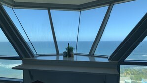 Et værelse med et stort karnapvindue med udsigt over havet, en lille plante på vindueskarmen og et skrivebord med en stol.
