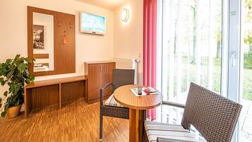 Ein Zimmer mit Holzboden, einem runden Tisch mit einer Zeitschrift, Korbstühlen, einem Holzschrank, einem Fernseher und einem Fenster mit Blick auf Grünflächen.