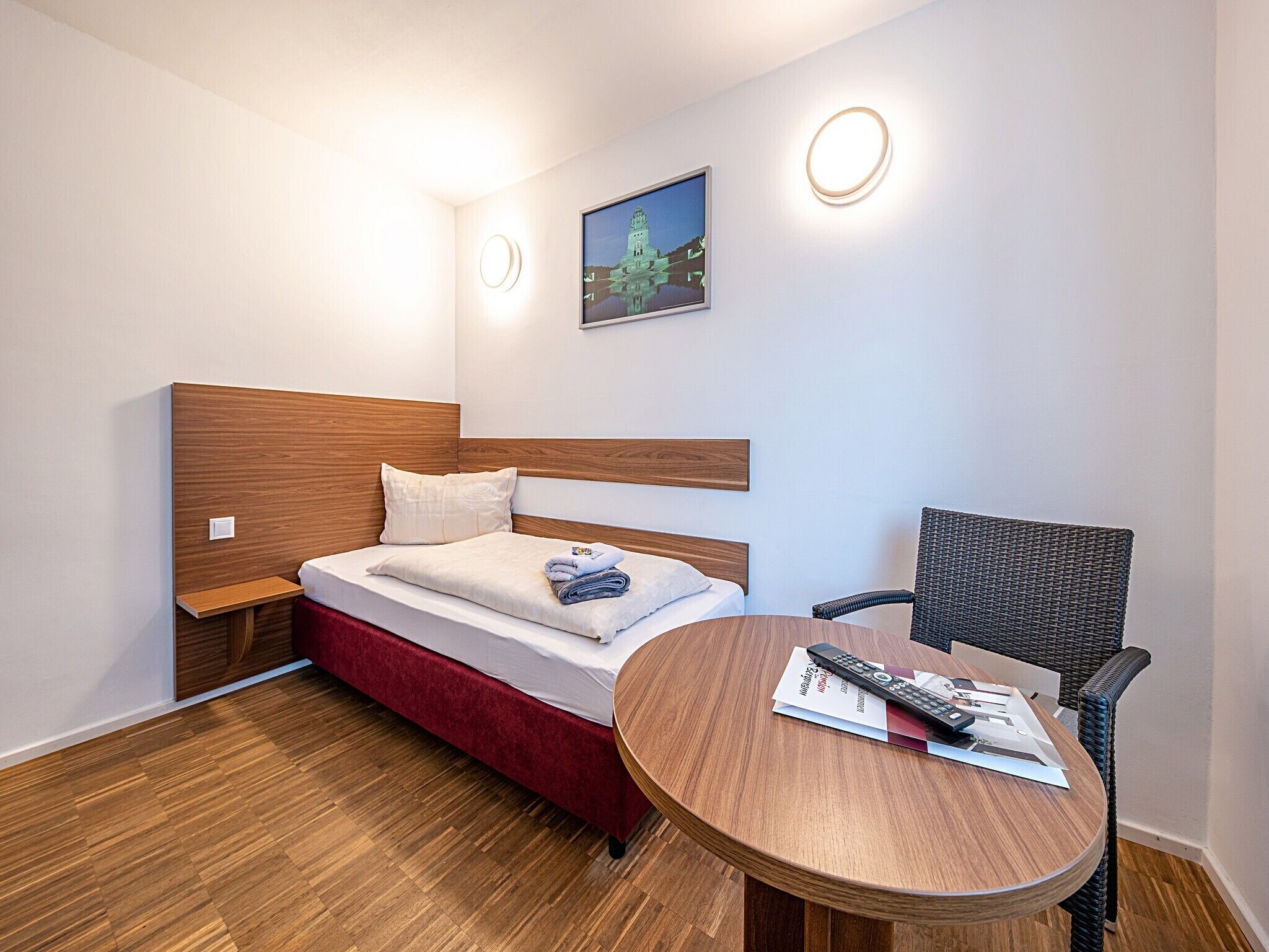 Ett hotellrum med en säng, ett runt bord i trä, en stol av rotting och en tavla som hänger på väggen.