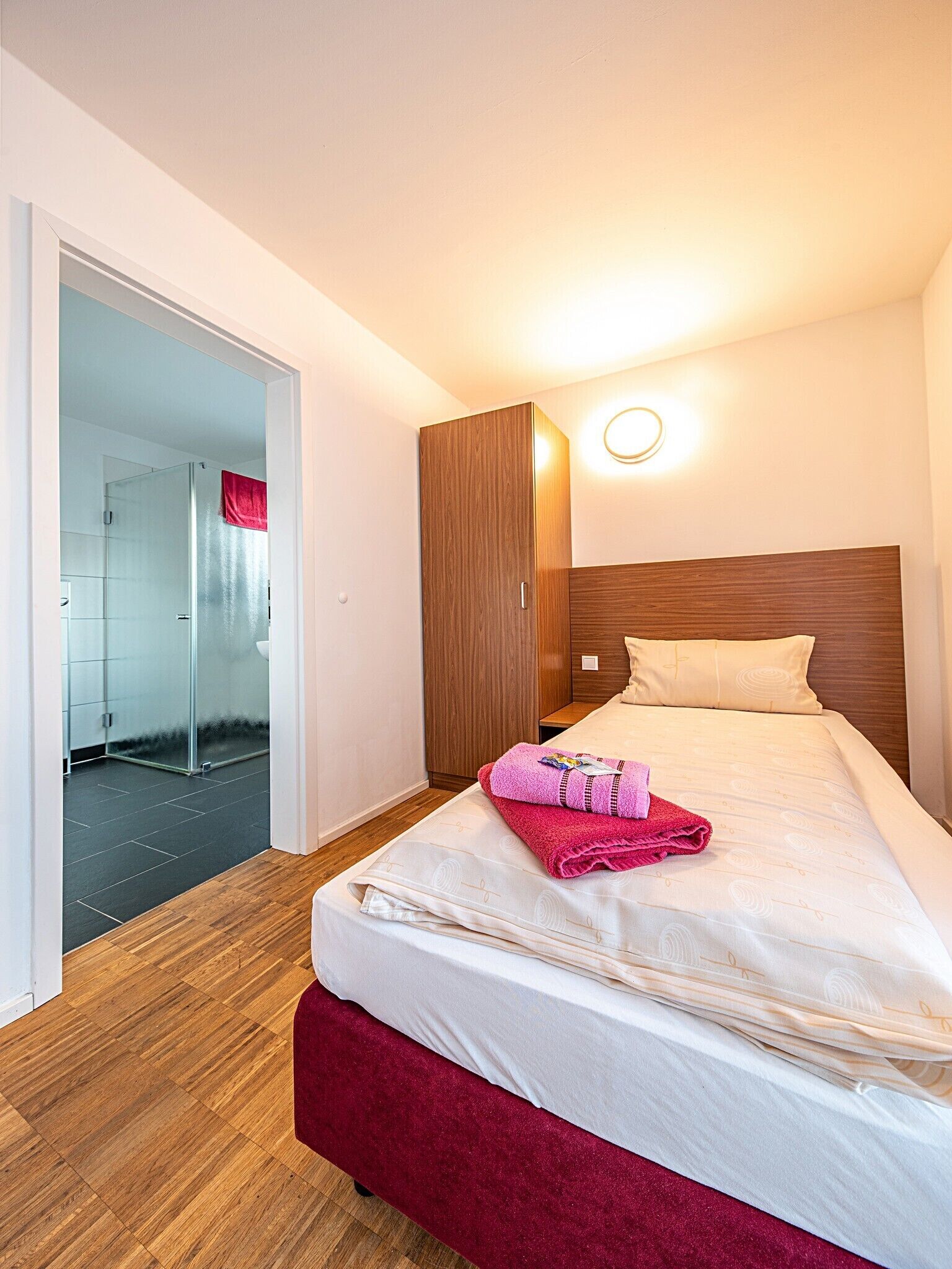 Hotellihuone, jossa on sänky, puinen vaatekaappi, lasinen suihkukaappi ja seinään kiinnitetty valaisin.