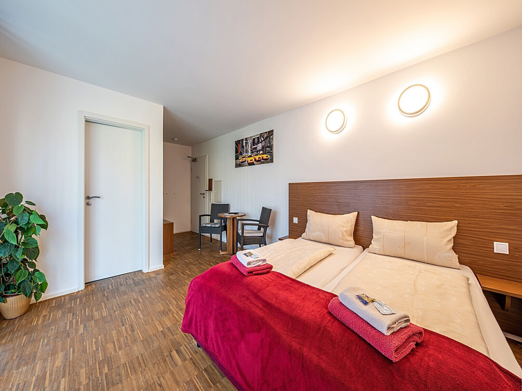 A hotel room with a wooden headboard, a red blanket, and a small desk with a chair.