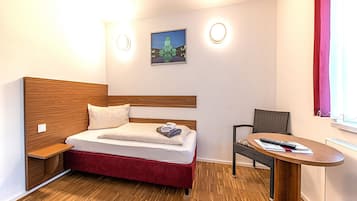 Habitación de hotel con cama de madera y cabecero, una mesita de noche pequeña, un sillón tapizado rojo, una mesa redonda de madera y un cuadro colgado en la pared.