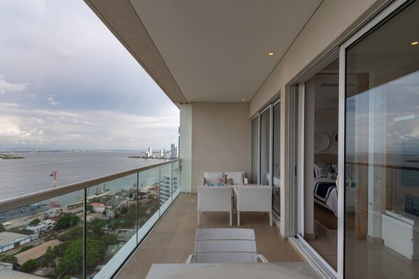 Varanda com vista para o mar e o horizonte da cidade, porta de vidro que dá para um quarto e uma pequena mesa com cadeiras.