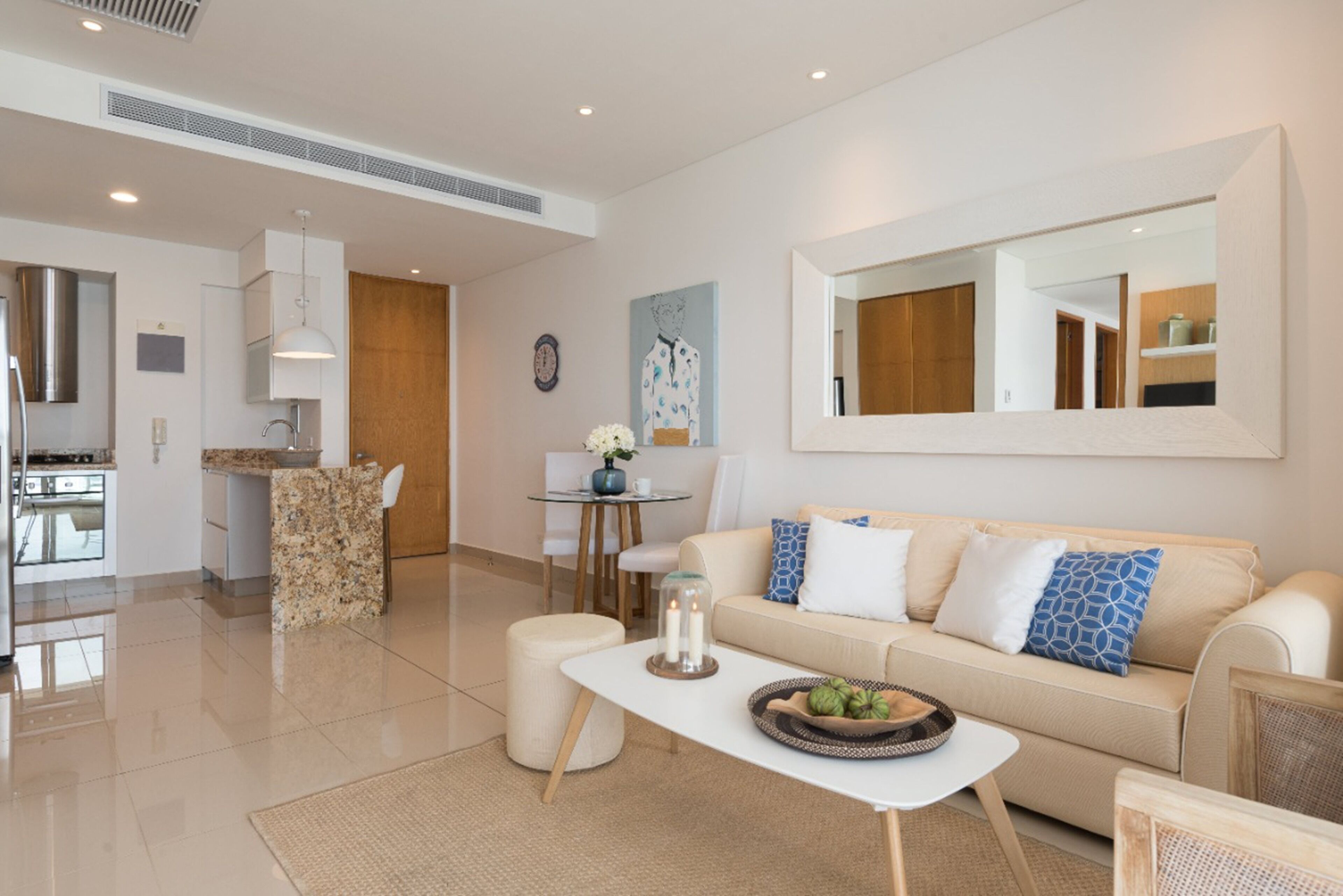 A modern living room with a white sofa, a coffee table with decorative items, a dining area with a small table and chairs, and a kitchen in the background.