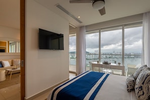 Quarto de hotel com uma cama grande, televisão de tela plana na parede, varanda com vista para a cidade e área de jantar com mesa e cadeiras.