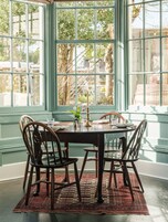 A dining area with a table set for four, surrounded by chairs, a patterned rug, and a window with a view of trees and buildings.
