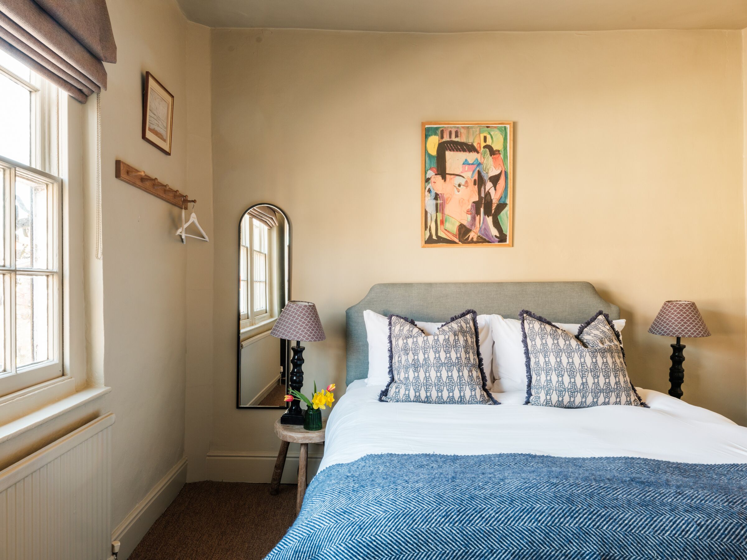 A bedroom with a bed, two lamps, a mirror, a picture on the wall, and a window.