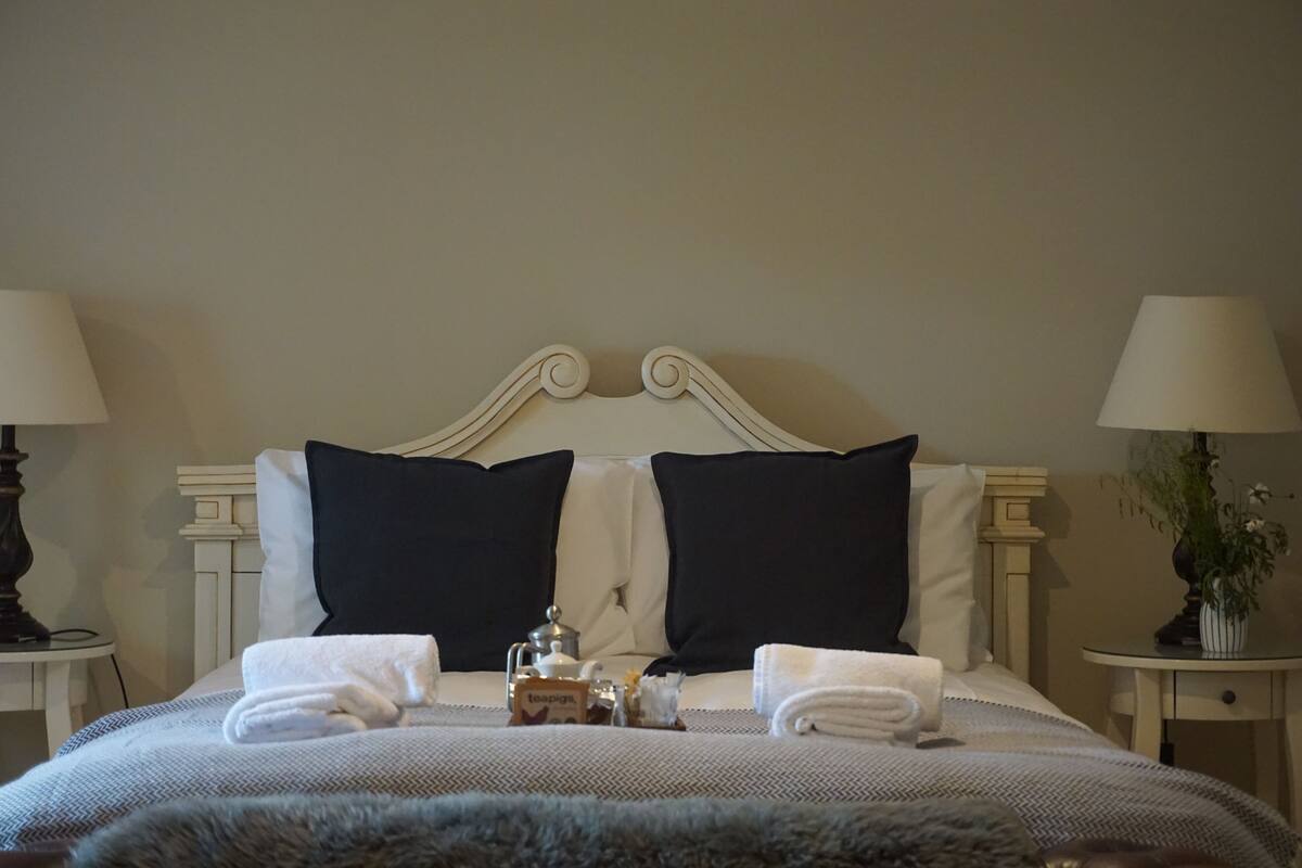A neatly made bed with two pillows, a bedside table with a lamp, and a small table with a plant and a teapot.