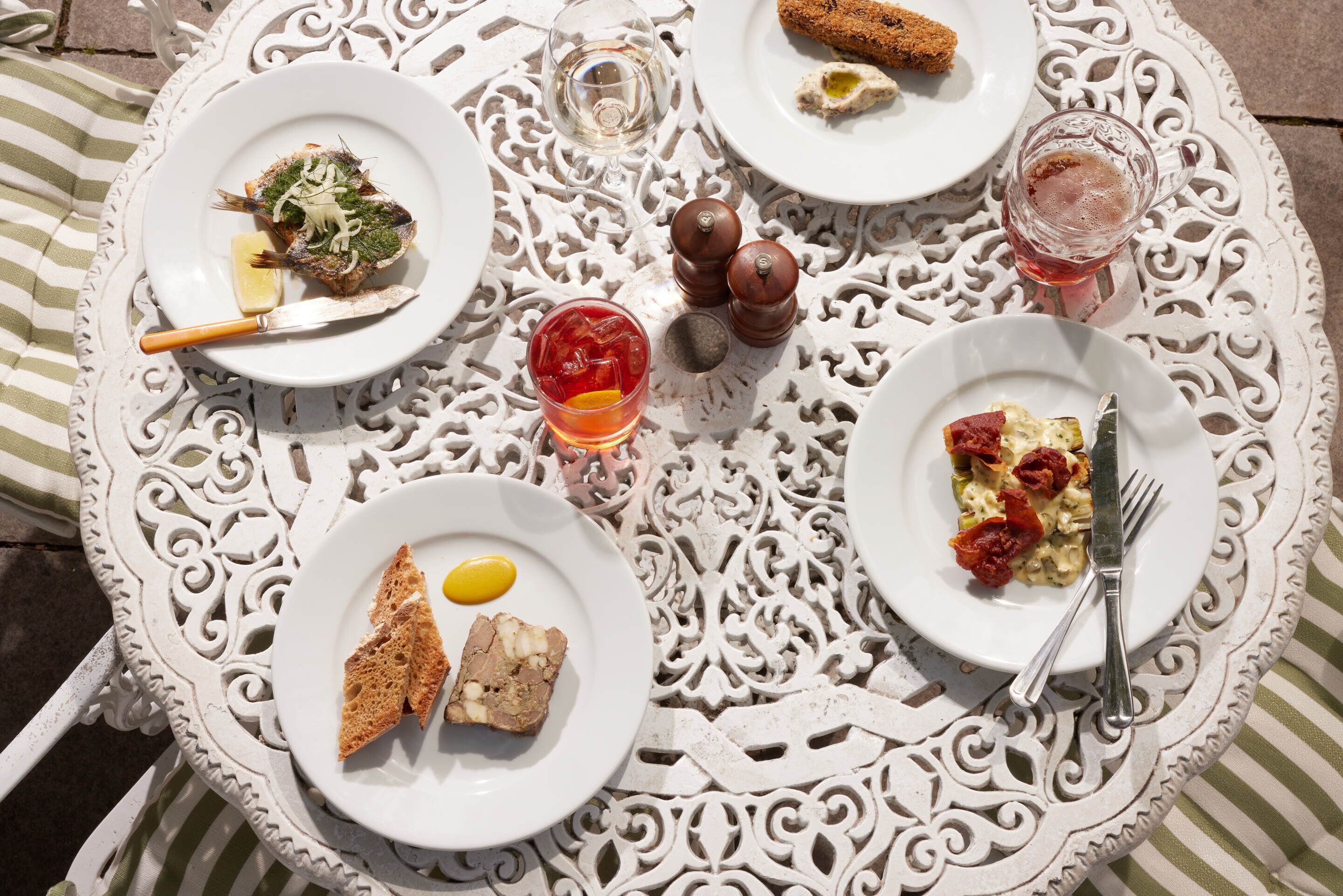 A round table with a white ornate design, surrounded by plates of food, cutlery, and condiments.