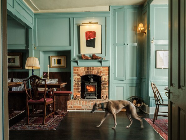 A room with teal walls, a brick fireplace, a dining table with chairs, and a dog walking through. - The Grosvenor Arms (Salisbury)