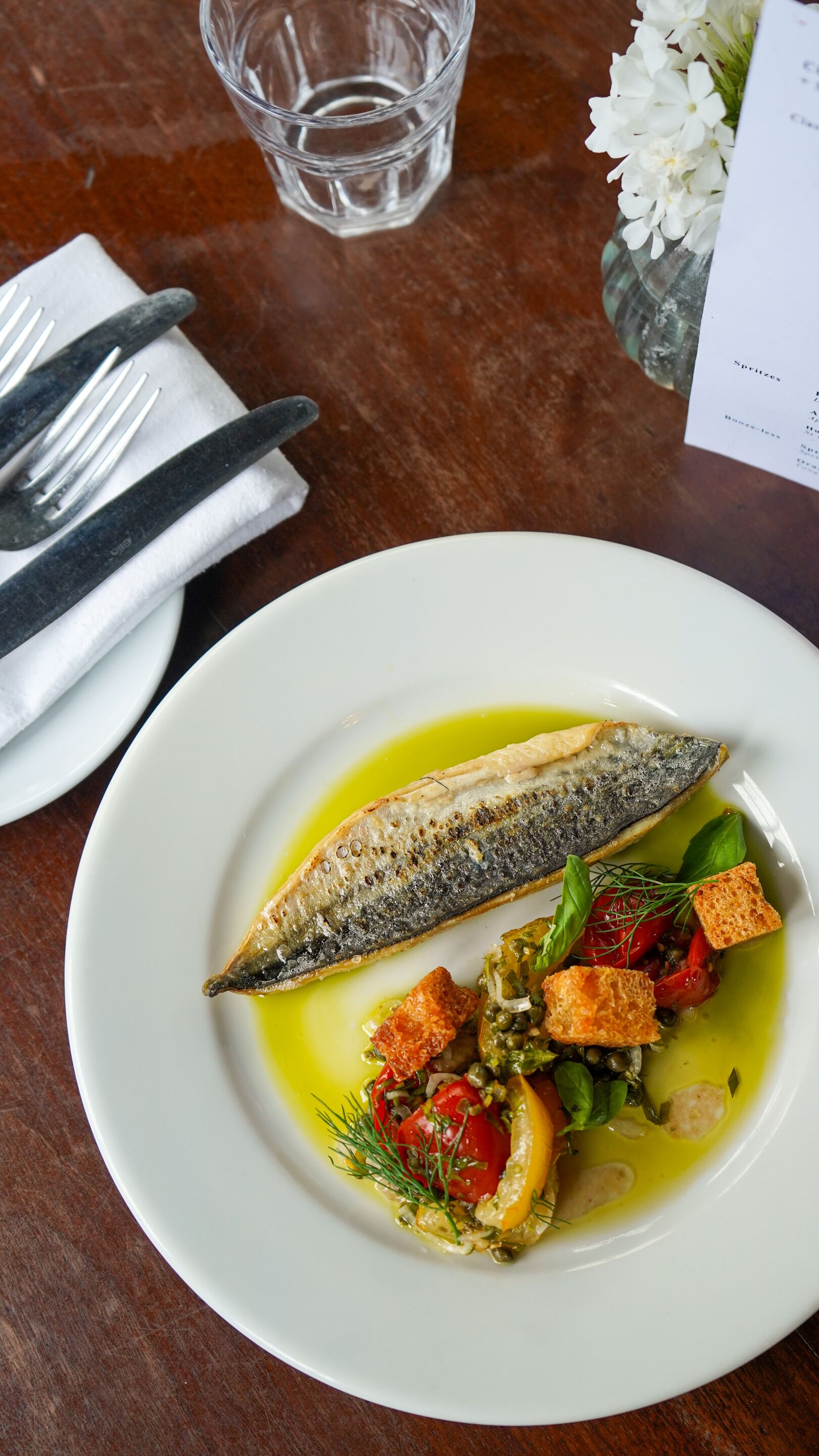 A plate of food with a fish, vegetables, and a glass of water on a wooden table.