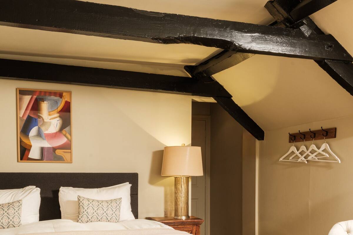 A bedroom with a large bed, a bedside table with a lamp, a chair, a coat rack, and a painting on the wall.