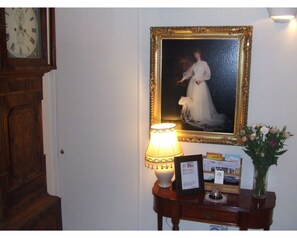 Een portret in een lijst aan de muur, een lamp op een tafel en een vaas met bloemen.