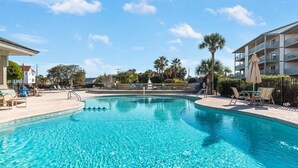 A clear swimming pool with lounge chairs and umbrellas, surrounded by palm trees and a building with balconies. - Lighthouse Point 25A | Ocean Views, Community Pool (Tybee Island)