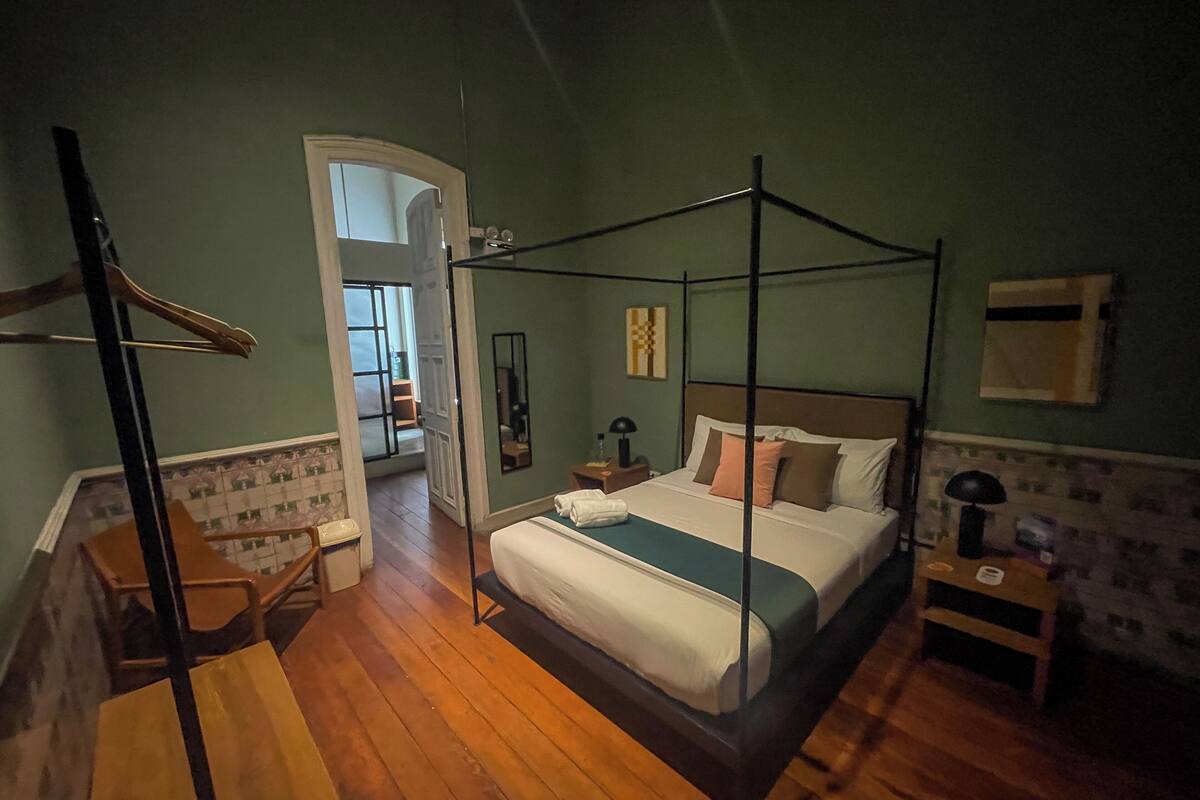 A four-poster bed with a canopy, a wooden floor, a small bedside table with a lamp, and a mirror on the wall.