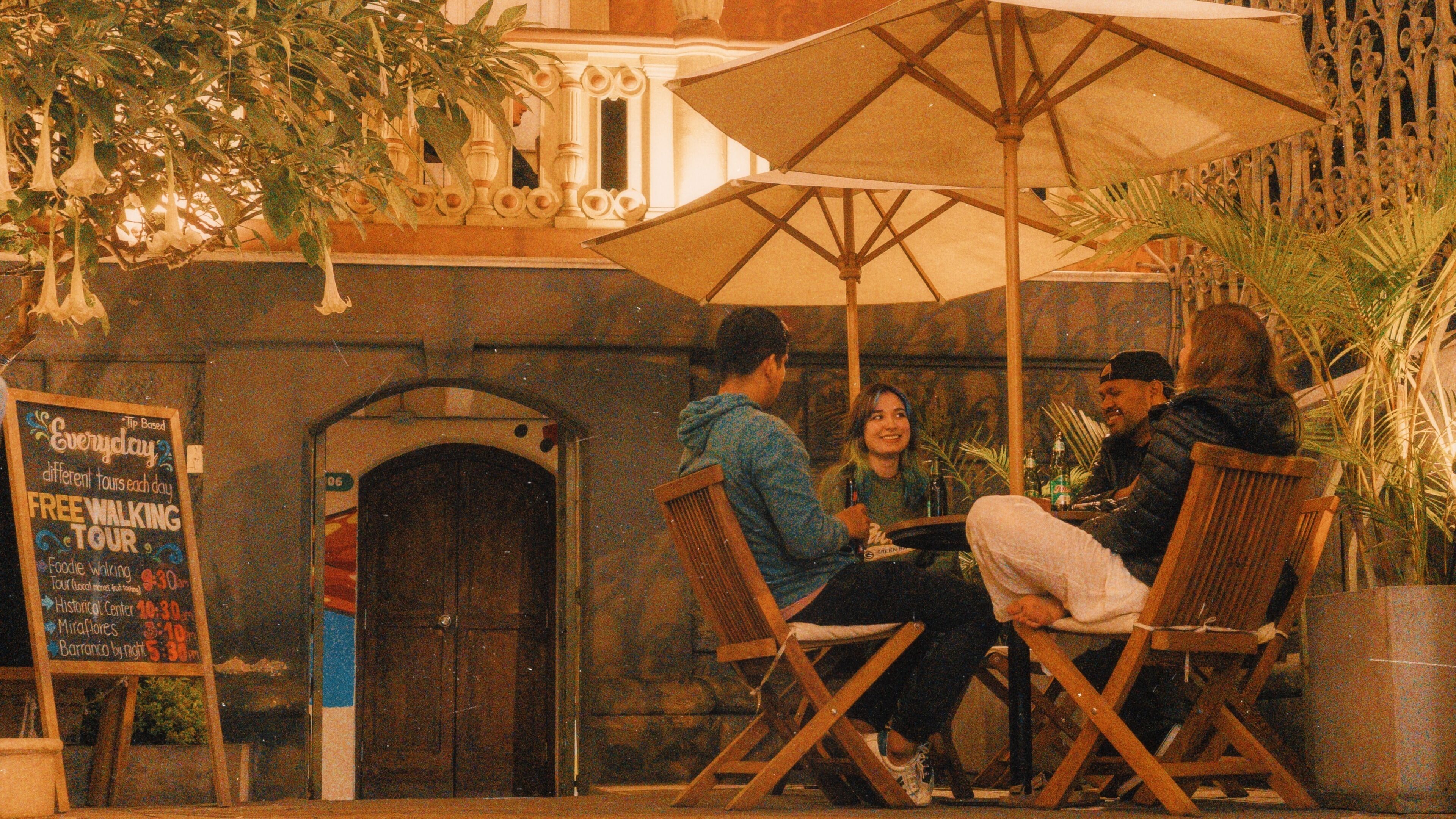 An outdoor seating area with wicker chairs and a large umbrella, a chalkboard sign with tour information, and a building with ornate details in the background.