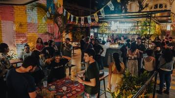 Un espacio interior concurrido, con mesas y sillas, gente sentada y de pie, y diversas plantas y adornos.