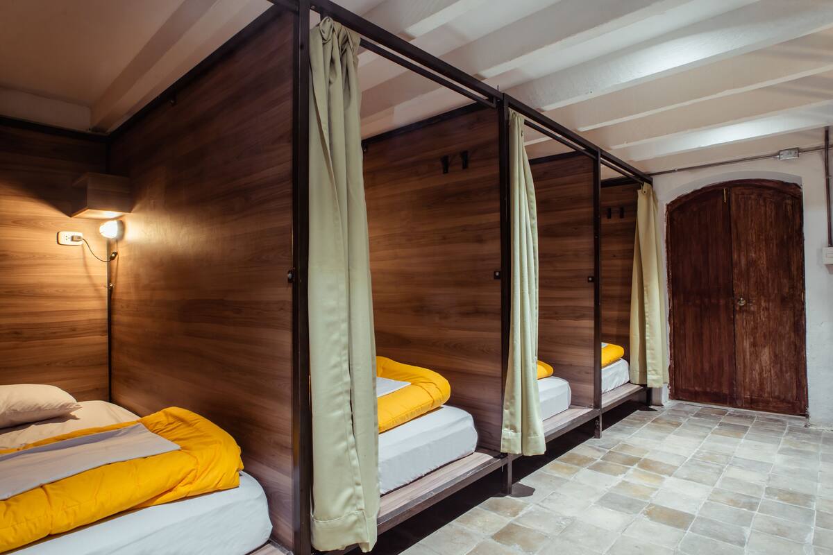 A room with four beds arranged in a row, each with a wooden headboard and a light fixture above.