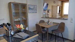 A dining area with a wooden table, two chairs, a kitchen counter with utensils, a microwave, and a cabinet with glassware.