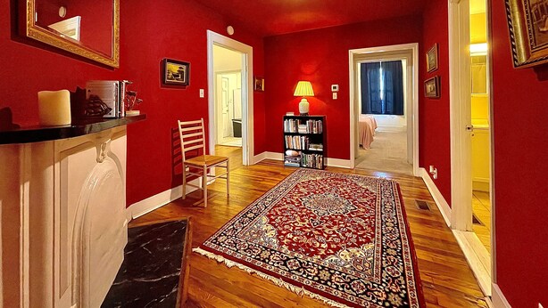 A hallway with red walls, a wooden floor, a large rug, a chair, a bookshelf, and a mirror.