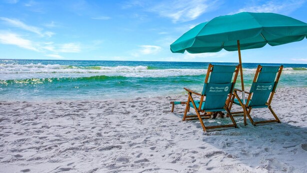 A beach with two beach chairs under a large umbrella, a sandy shore, and the ocean in the background.