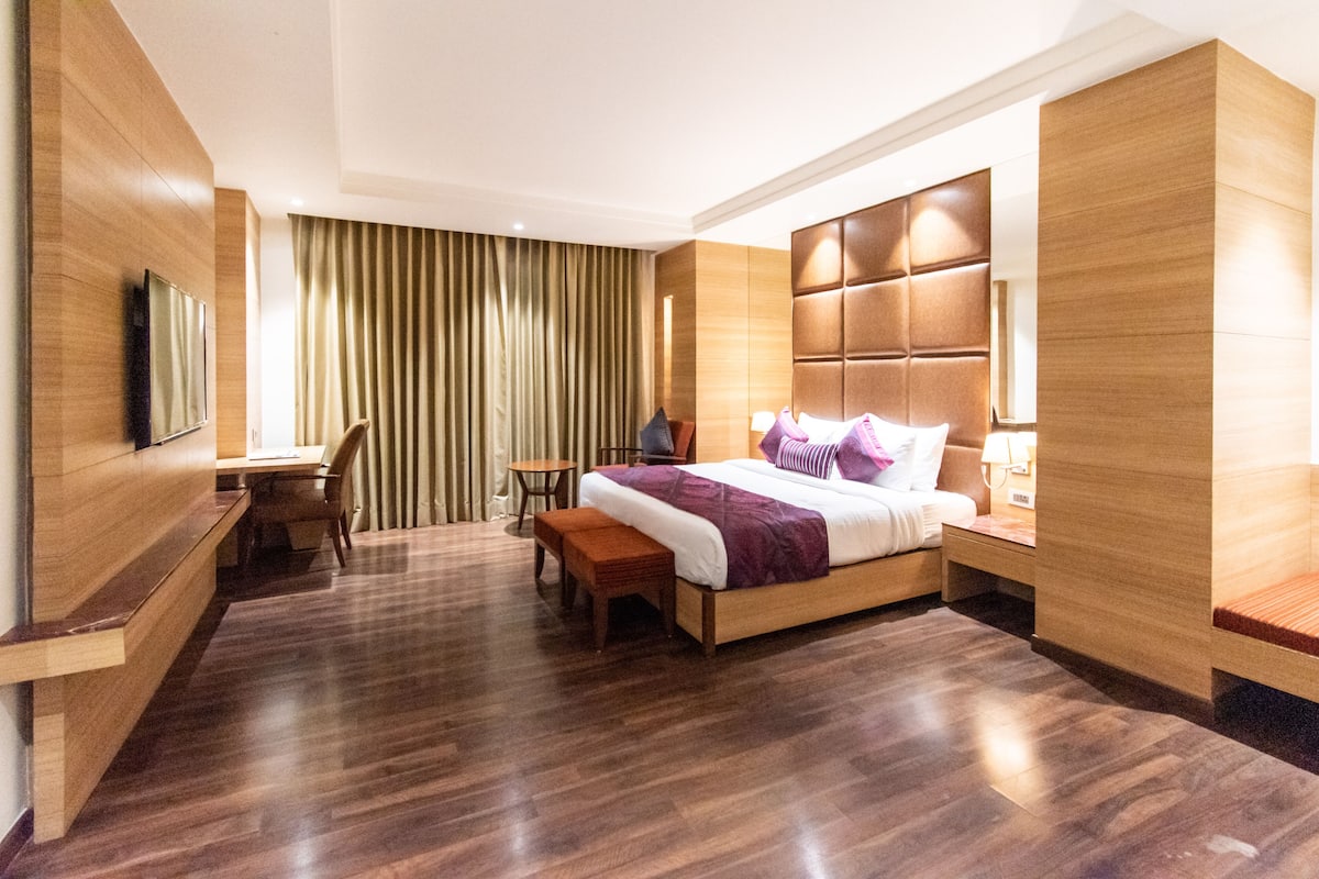 A modern hotel room with a wooden floor, a large bed with a headboard, a desk with a chair, a bench, and a TV mounted on the wall.