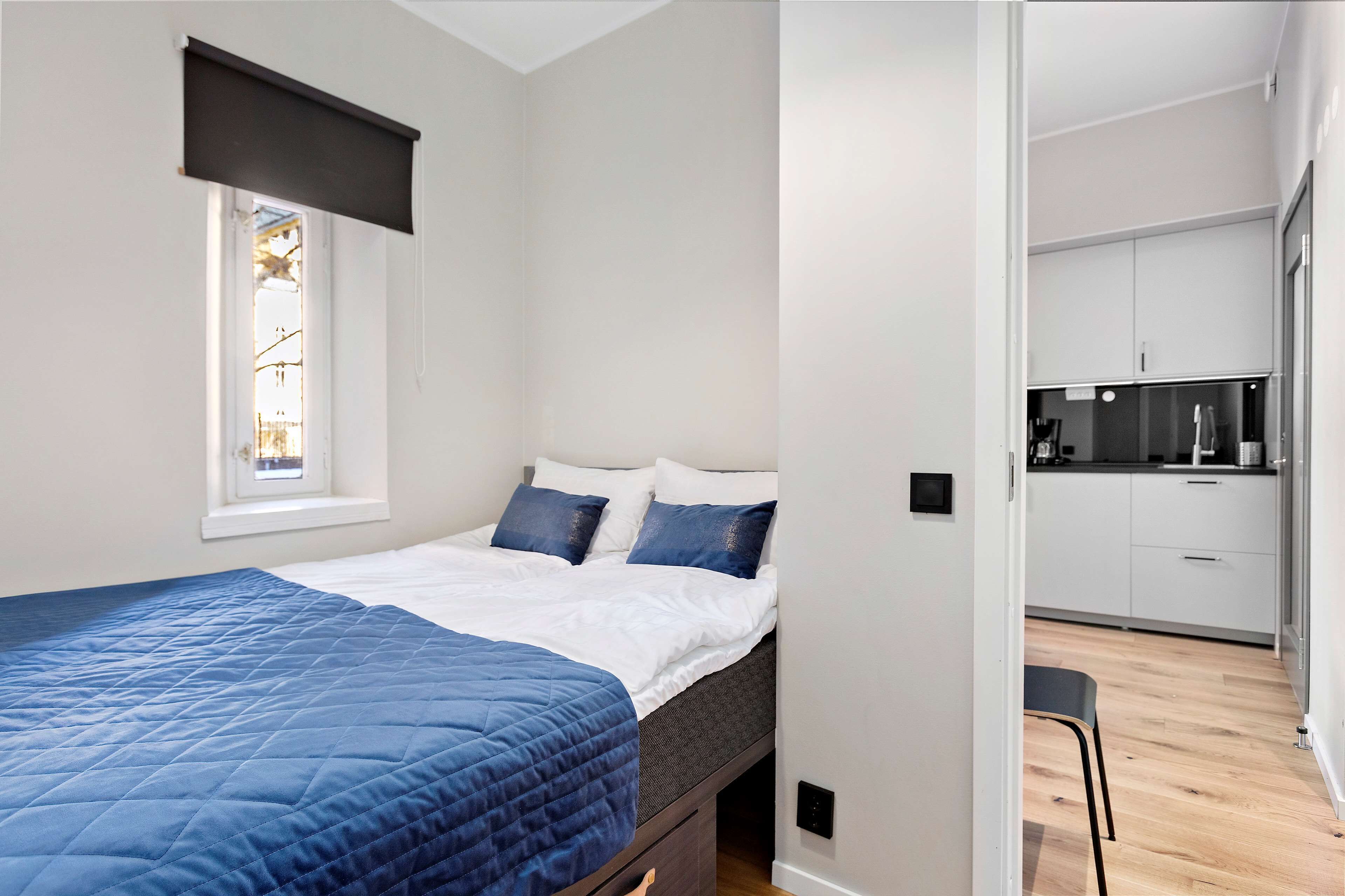 A bedroom with a bed, a window, and a view into a kitchen area.