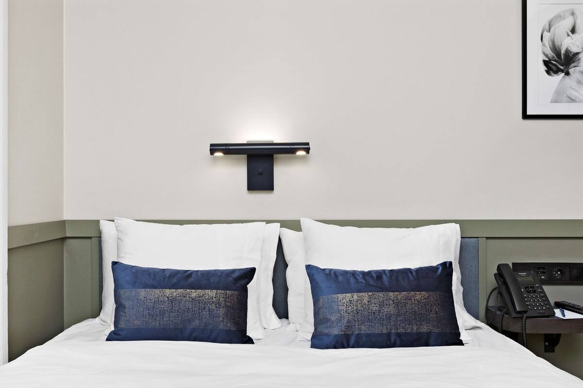 Habitación de hotel con cama, almohadas, mesita de noche con teléfono y lámpara de pared.
