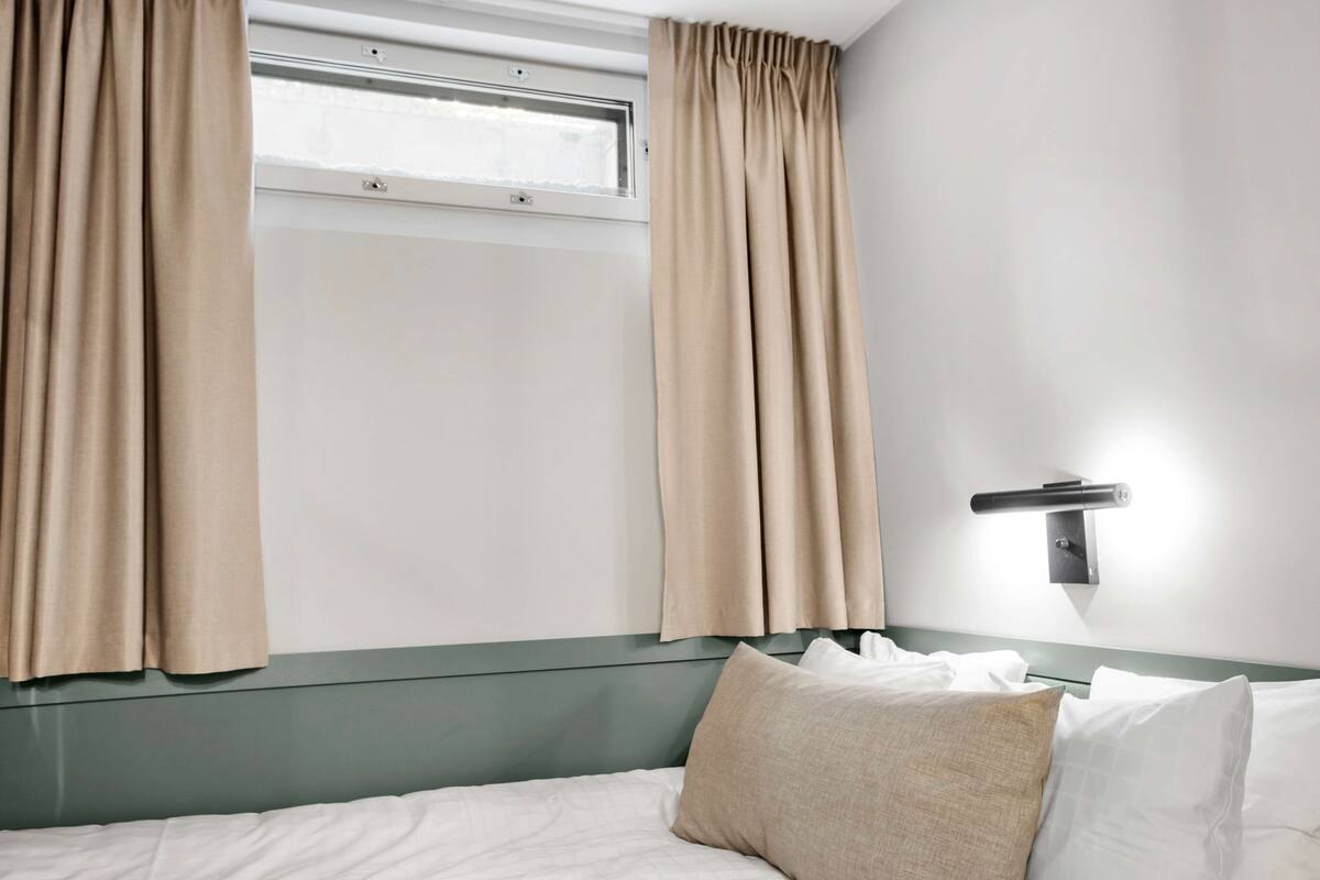 Habitación de hotel con cama, cortinas color beige, ventana y lámpara fijada a la pared.