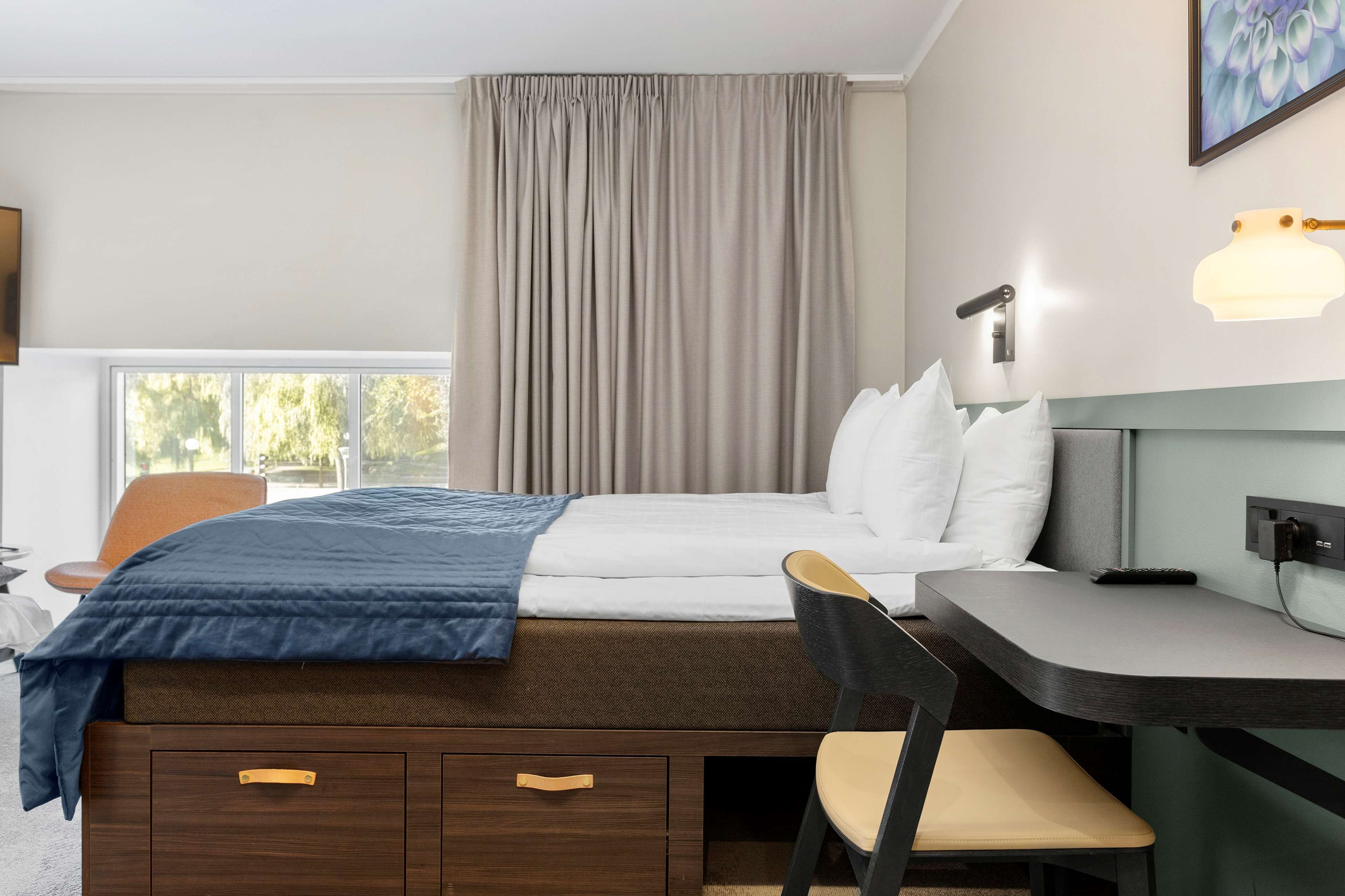 A modern hotel room with a bed, desk, chair, and a large window.