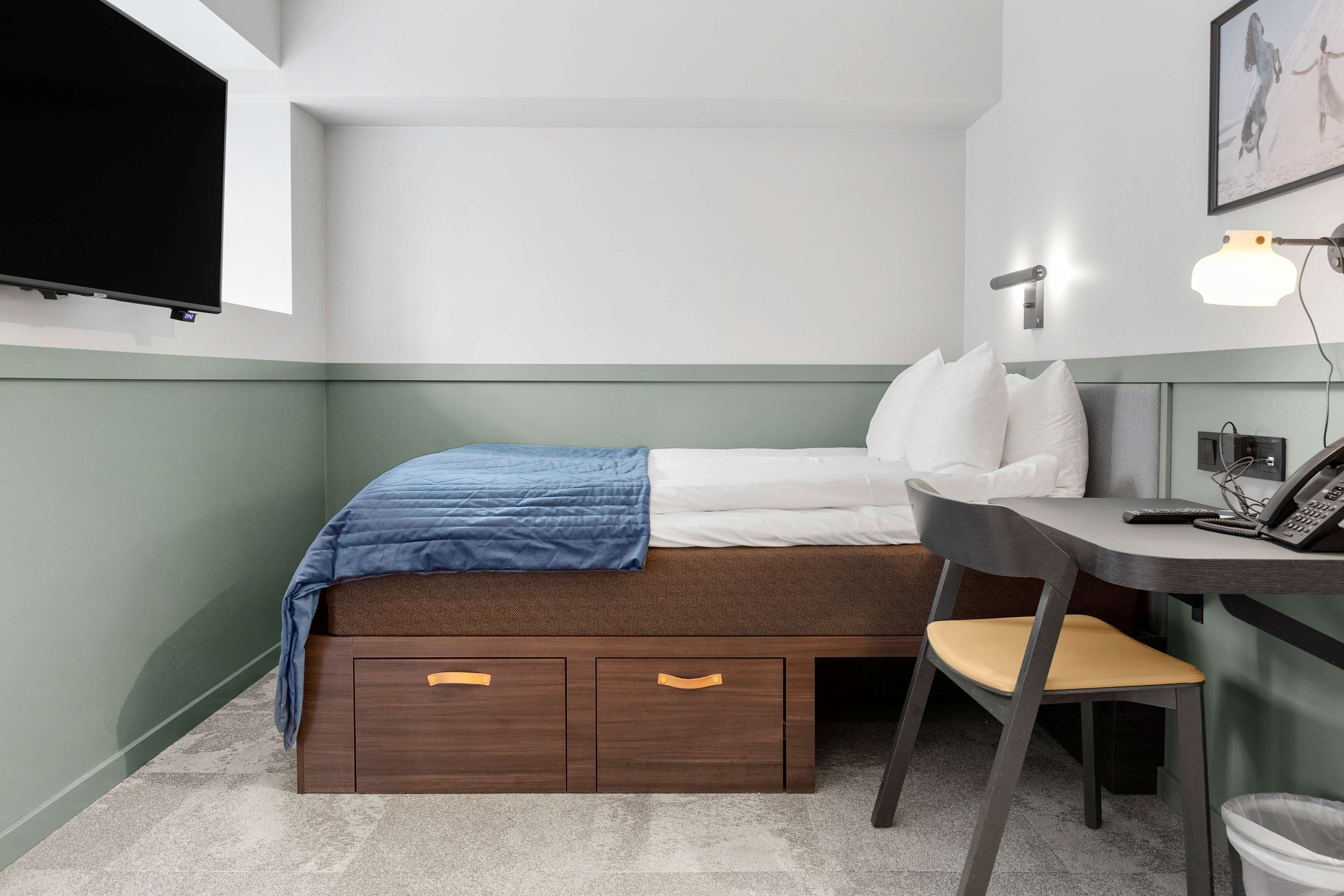 A compact hotel room with a bed, a desk with a chair, a desk lamp, a mounted TV, and a framed picture on the wall.