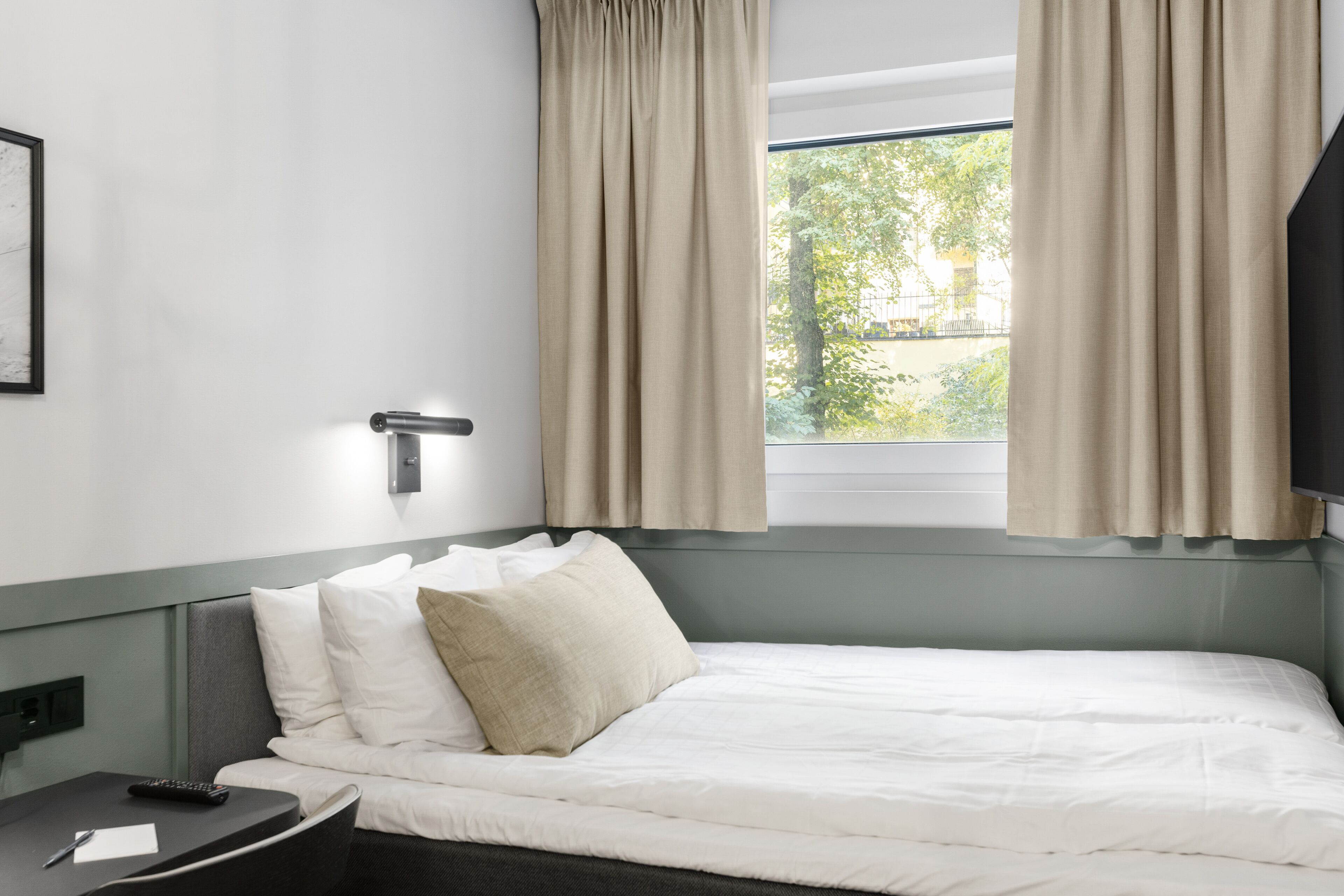 Una habitación de hotel con una cama, una mesita de noche con un control remoto, una ventana con cortinas y una lámpara fijada a la pared.