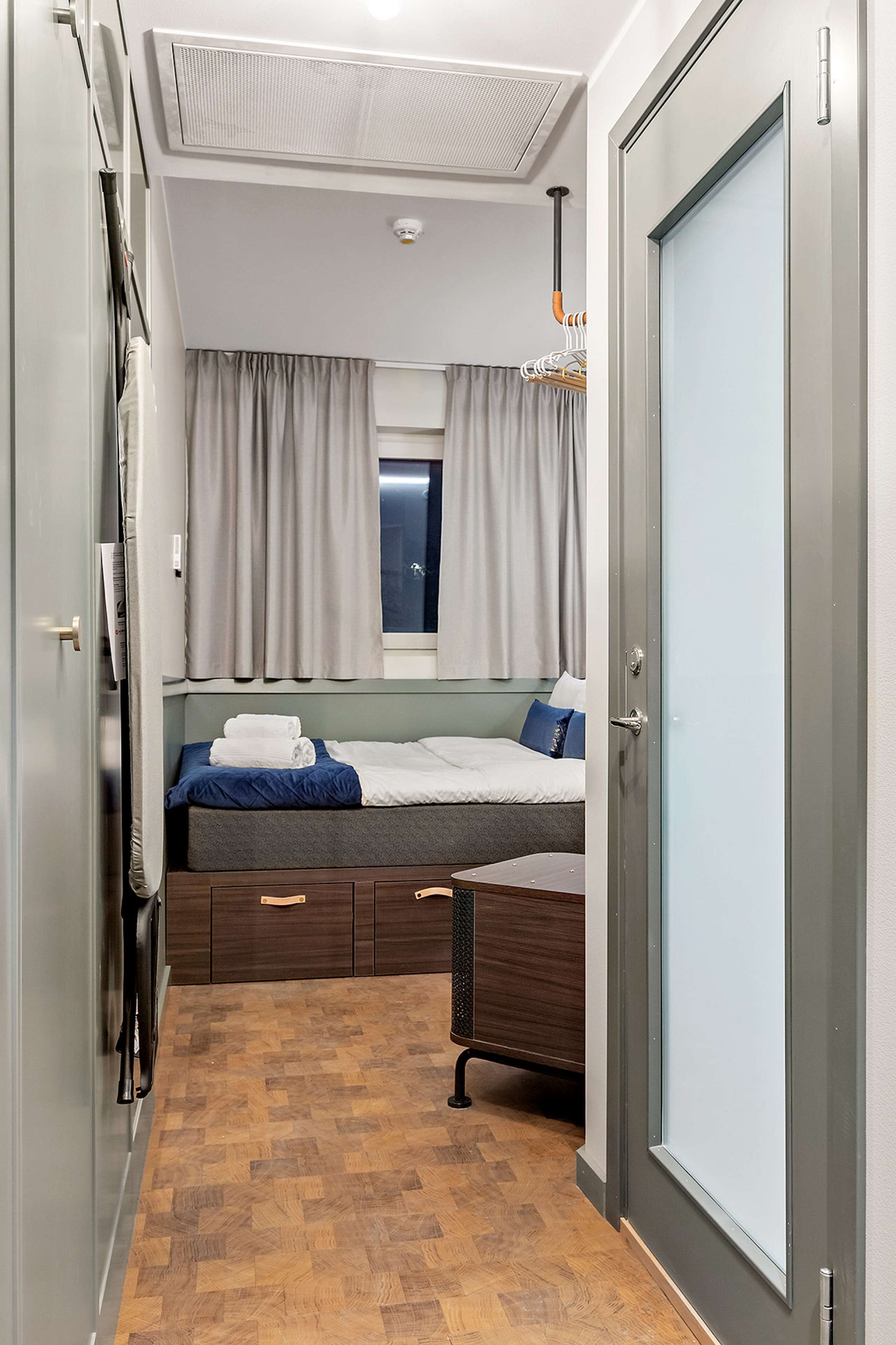 A compact hotel room with a bed, a small desk, a window with curtains, a ceiling fan, and a door.