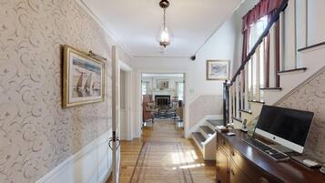 A hallway with patterned wallpaper, a framed painting, a staircase with a red curtain, and a desk with a computer and keyboard.
