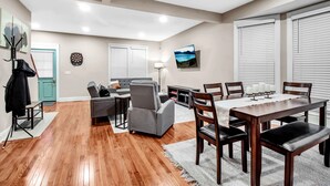 A spacious living area with a grey sofa, a wooden dining table with six chairs, and a flat-screen TV mounted on the wall.