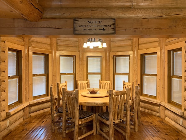 Cozy cabin-style room with a central dining table and six chairs, large windows, and exposed wooden beams.