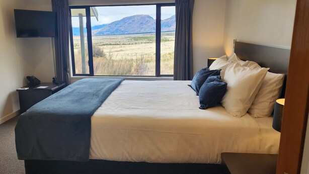 A bedroom with a large bed, a flat-screen TV, a bedside table, and a window with a mountain view.