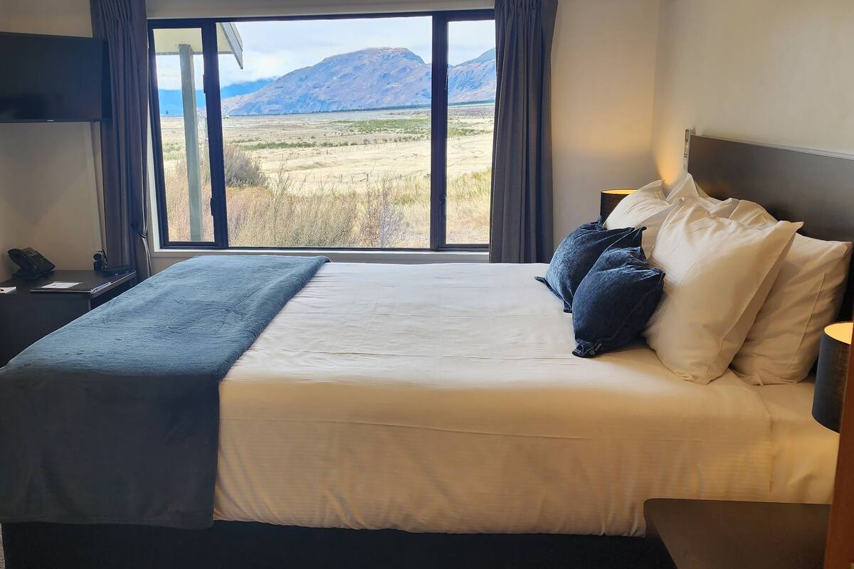 Un dormitorio con una cama grande, un televisor de pantalla plana, una mesita de noche y una ventana con vista a la montaña.
