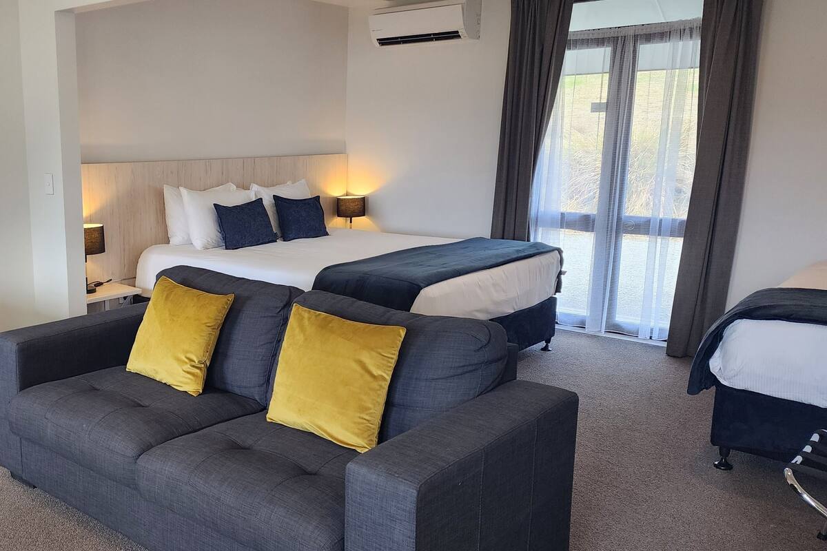 Habitación de hotel con una cama grande, un sofá y una cama individual. Hay un cuadro en la pared y una ventana con cortinas.