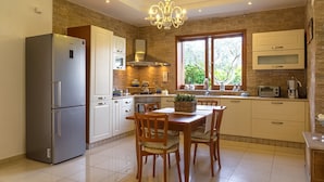 Eine moderne Küche mit einem Edelstahl-Kühlschrank, einem Holzespeisetisch und Stühlen. Eine Kristallleuchter hängt von der Decke und ein Fenster bietet Blick auf Grünfläche im Freien.