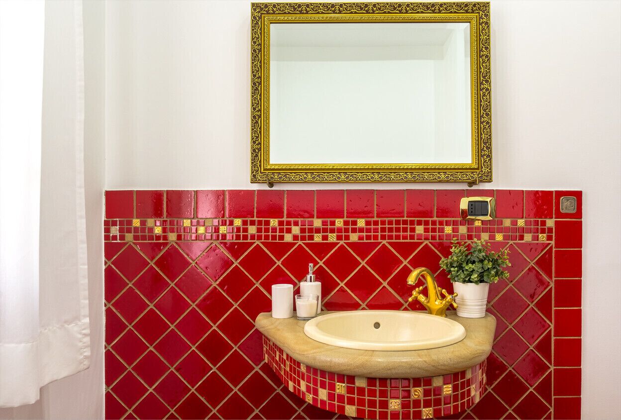 Een badkamer met een rode betegelde spatwand, een spiegel met gouden lijst, een wastafel met gouden kraan en een potplant.