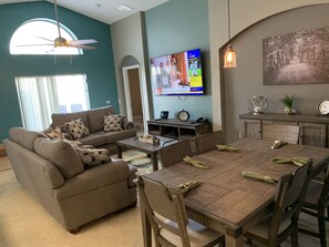 A living room with a sofa, coffee table, dining table, chairs, a TV, and a painting on the wall. - 4 BED 3 BATH SUN KISSED LUXURY VILLA (Kissimmee)