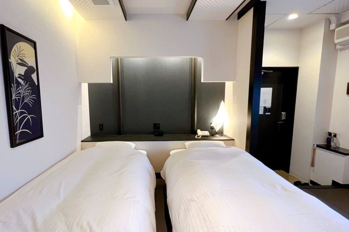 A hotel room with two single beds, a large mirror, a bedside table with a lamp, and a framed artwork on the wall.